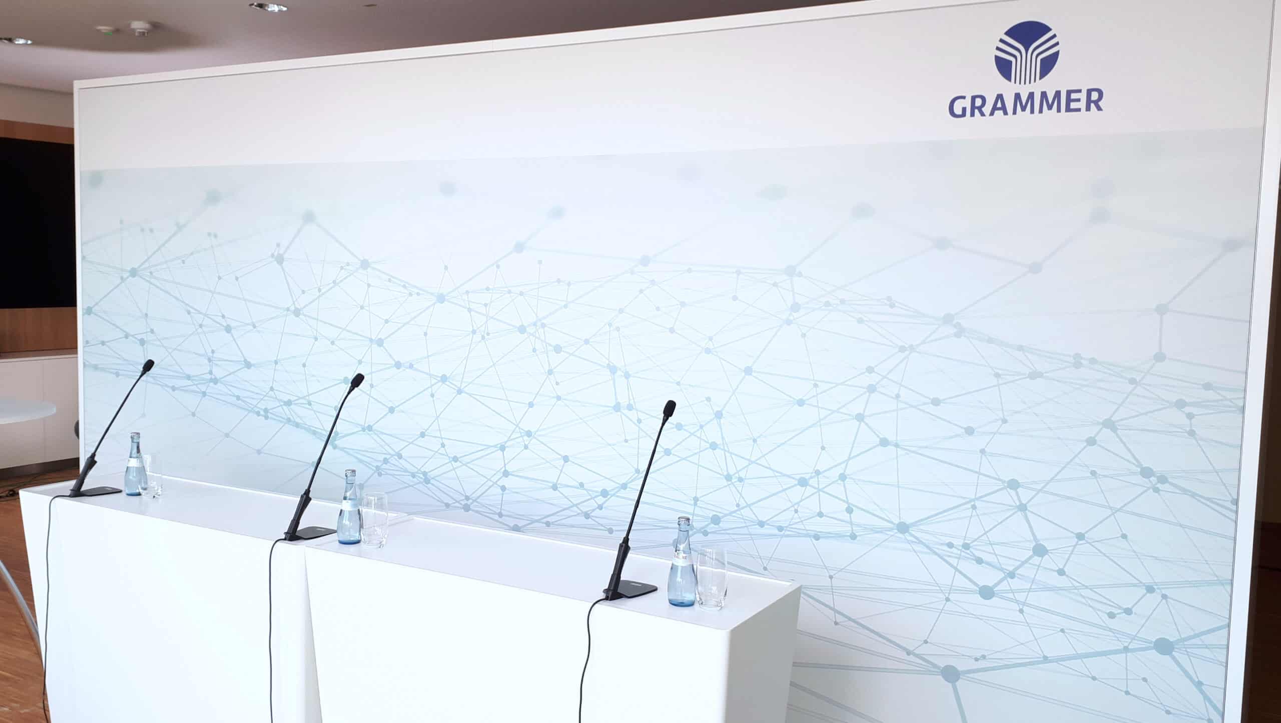 Konferenztisch mit drei Mikrofonen und Wasserflaschen vor GRAMMER Logo und Netzwerk-Grafik