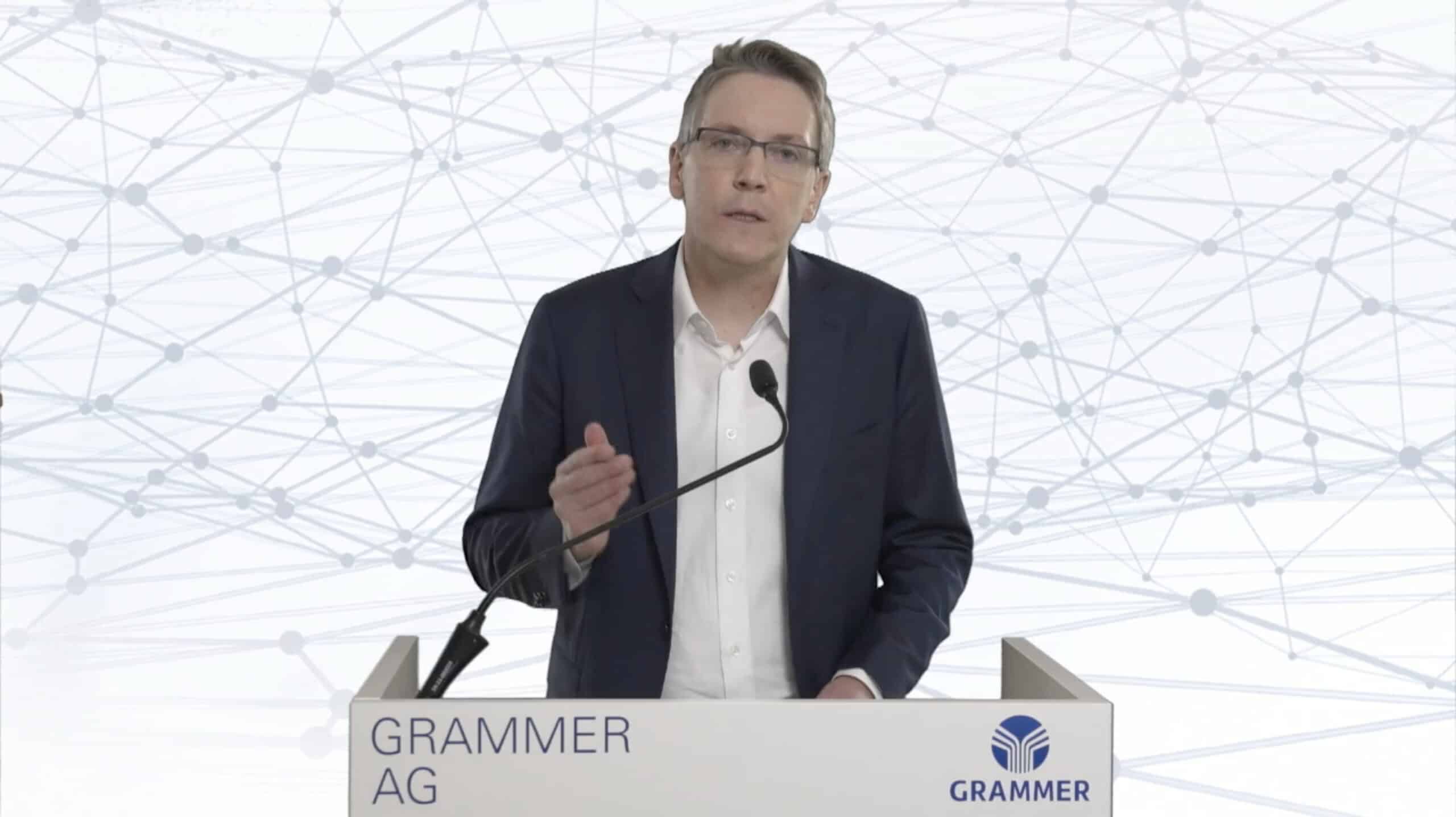Geschäftsmann in Anzug spricht an Grammer AG Pult vor digitalem Netzwerk-Hintergrund