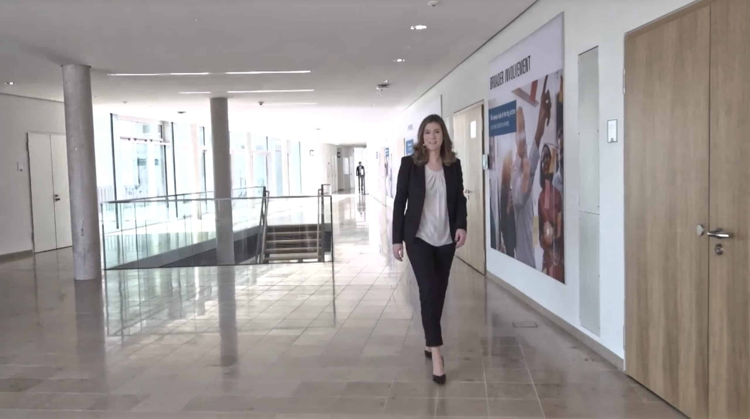 Businessfrau läuft lächelnd durch modernen Flur mit Diversity-Poster 'Broader Involvement'