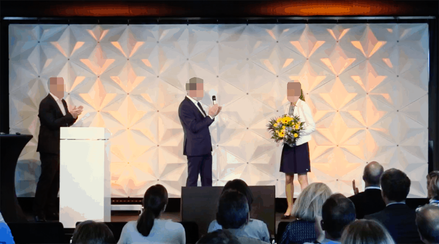 Bühnenmoment bei internem Livestream-Event: Frau mit Blumenstrauß, zwei Männer applaudieren vor geometrischem Hintergrund