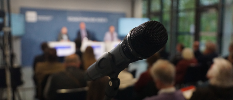 Nahaufnahme eines Shure-Mikrofons bei einer Vorwerk-Konferenz mit Bühne und Publikum im Hintergrund