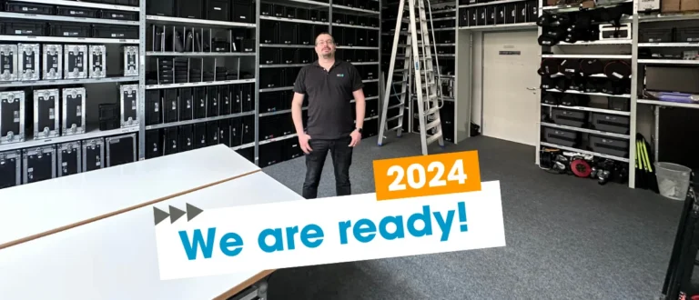 NC3 Mitarbeiter in Technik-Lager mit Event-Equipment und 'We are ready! 2024' Banner