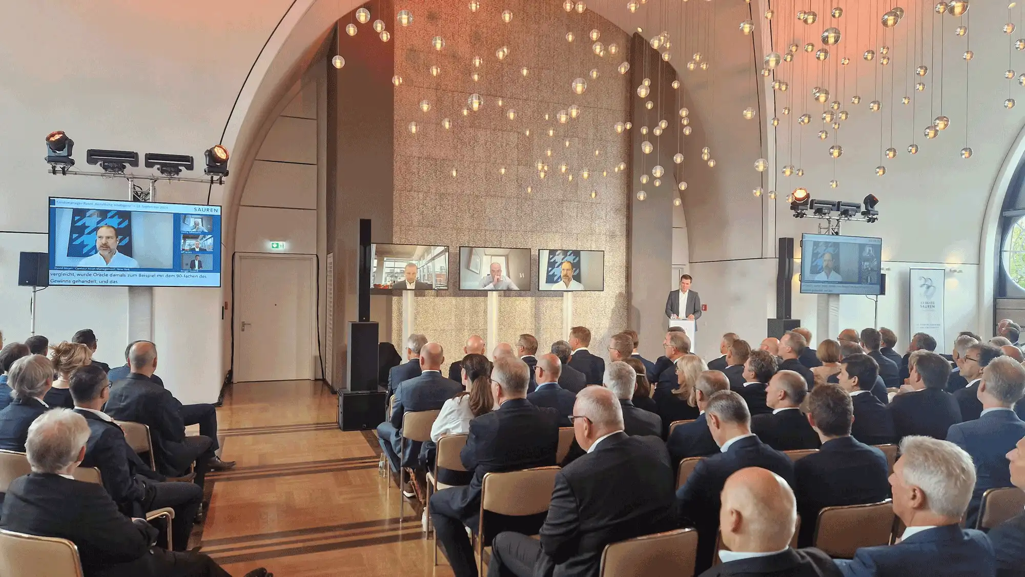 event-location-für-ki-panel Hybrid-KI-Panel in moderner Event-Location mit Publikum, Bühne und Remote-Speakern auf Großbildschirmen