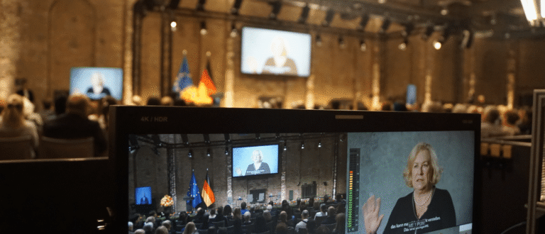 Monitor zeigt Livestream einer Gedenkstunde mit Rednerin, Publikum und Deutschland- sowie EU-Flaggen im Saal