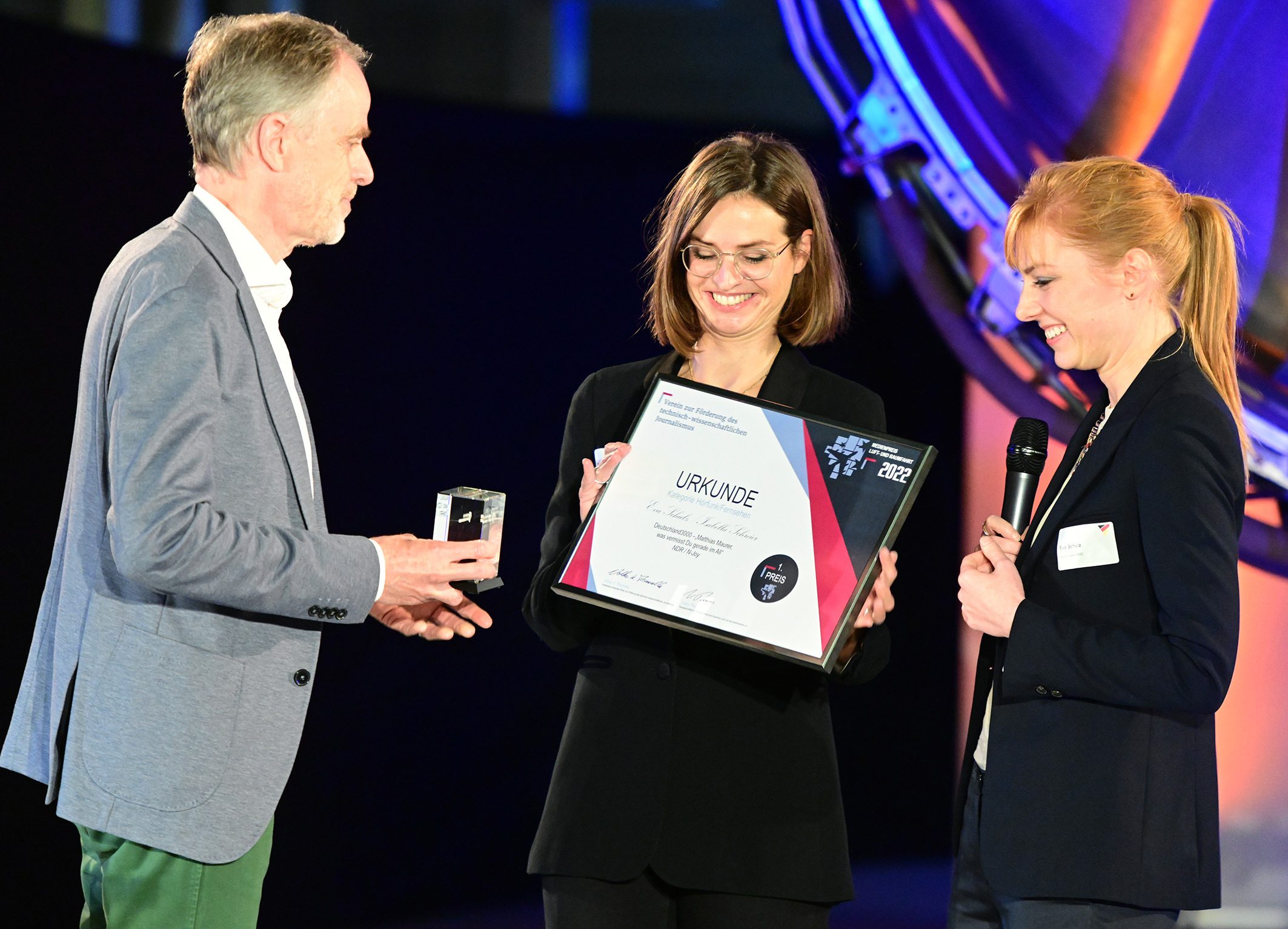 Preisverleihung Medienpreis LfK 2022: Übergabe von Urkunde und Pokal auf Bühne mit Moderatorin
