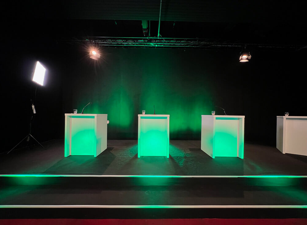 verbio-hv-studio2-ansicht-veranstaltung-live-streamen.jpg Bühnenaufbau mit vier weißen Pulten, grüner LED-Beleuchtung und professioneller Lichttechnik für Livestream im HV Studio2