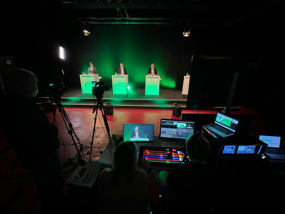 verbio-hv-studio4-ansicht-veranstaltung-live-streamen.jpg Blick in Verbio HV Studio 4: Bühne mit drei Pulten, grüner Beleuchtung und Streaming-Regie mit Monitoren und Kameras