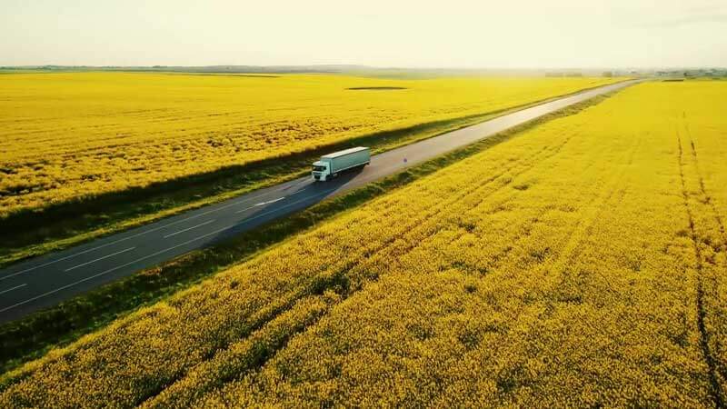 Drohnenaufnahme eines weißen LKWs auf Landstraße durch weite gelbe Rapsfelder bei Sonnenlicht