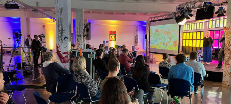 Videoproduktion kidsCon 2022: Kameras, Bühne mit Sprecher, großem Screen und Publikum in farbig beleuchteter Halle